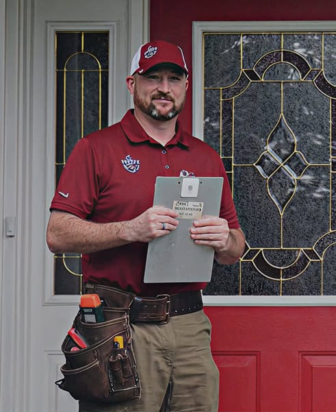 A man in a red polo shirt and cap stands in front of a red door, holding a clipboard. Wearing a tool belt with various tools, he represents Anchor Home Inspection, ready to provide thorough CT Home Inspection services.