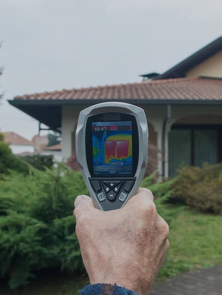 A person holds a thermal imaging camera pointed at a house, displaying a heat map of the building on the device's screen during an Anchor Home Inspection.