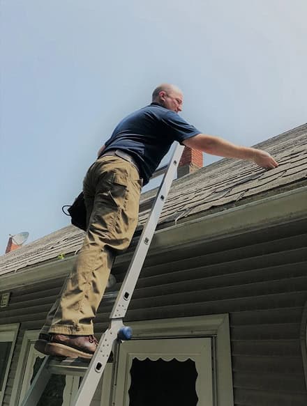 A man stands on a ladder, inspecting or working on the roof of a house under daylight—an expert approach you can expect from Anchor Home Inspection.