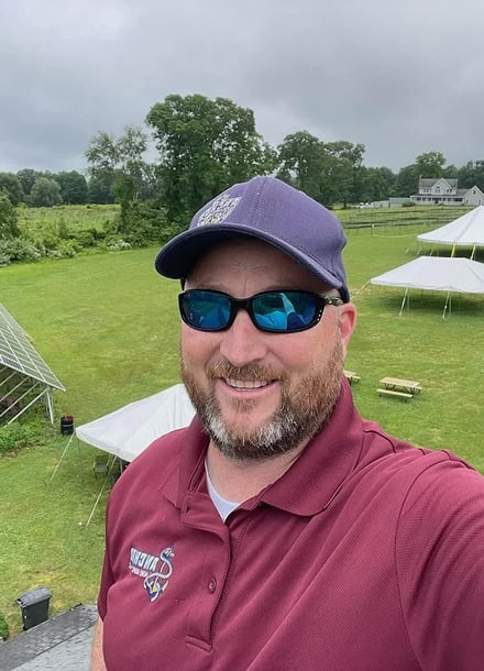Man wearing sunglasses, a navy cap, and a maroon polo shirt stands outdoors, smiling, with tents and green fields in the background under a cloudy sky—an inviting scene at an Anchor Home Inspection event.