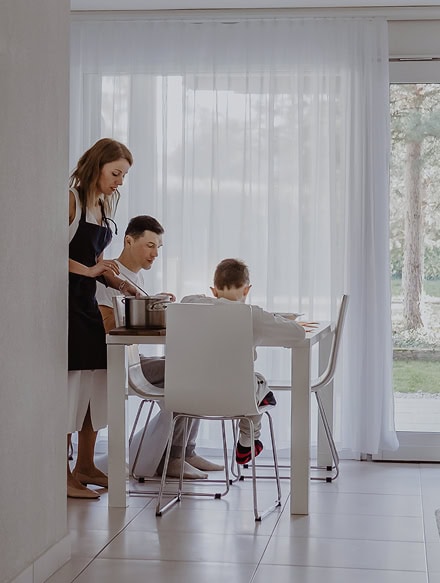 A woman serves food at a kitchen counter while a man and child sit at a dining table in a bright room with large windows, reflecting the comfort and safety you expect after an Anchor Home Inspection.